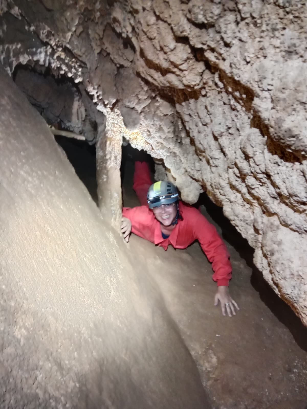 Smiling caver crawling through a low rocky passage inside Cueva Forat la Bou wearing a red protective suit, helmet and headlamp