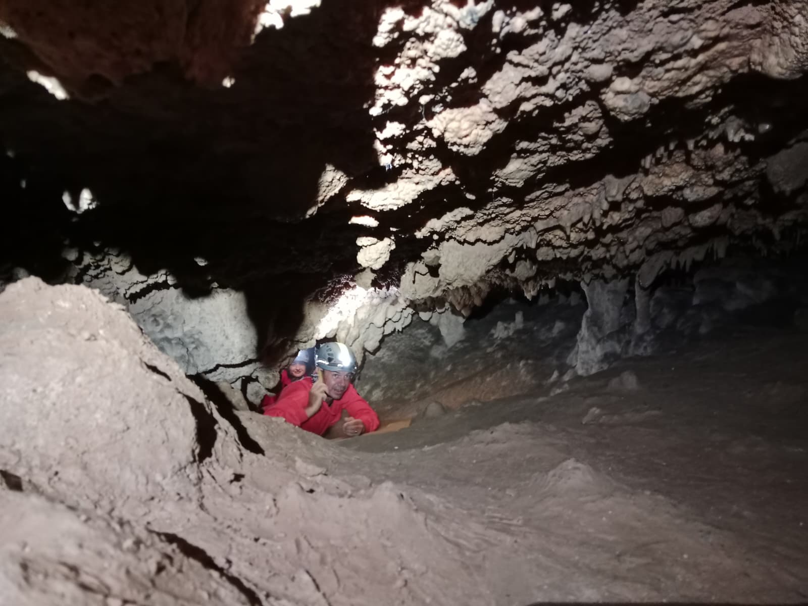 Two cavers crawling through a low passage inside Cueva Forat la Bou, wearing red protective suits and helmets beneath striking layered cave formations
