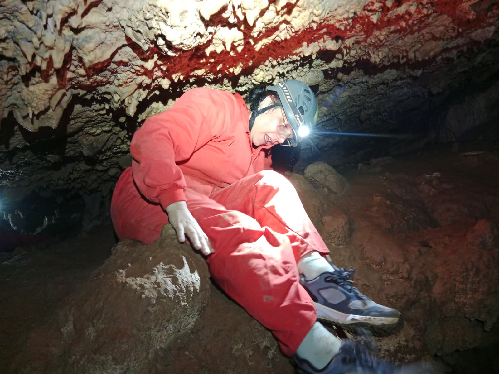 Caver moving through a low passage inside Cueva Forat la Bou, wearing a red protective suit and helmet with headlamp beneath striking mineral formations