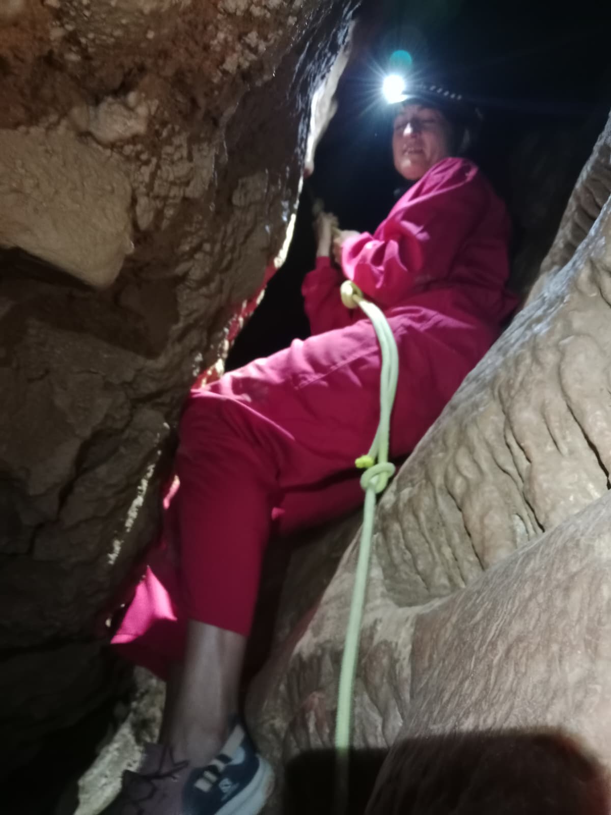 Caver descending through a narrow passage inside Cueva Forat la Bou, wearing a pink protective suit and helmet with headlamp while using a rope
