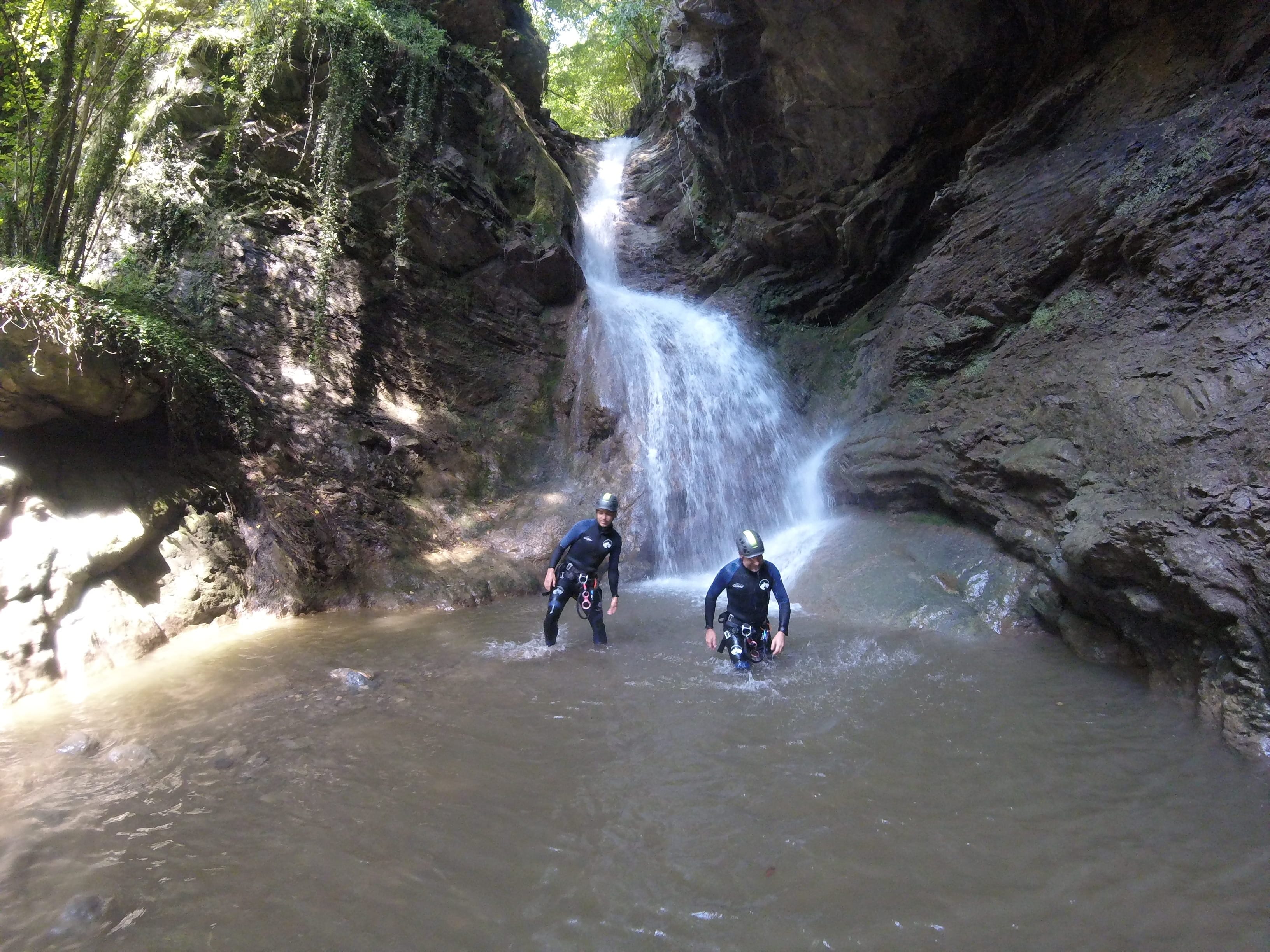 Dos participants de barranquisme drets en una poza sota una gran cascada al bosc del Barranc del Pamano